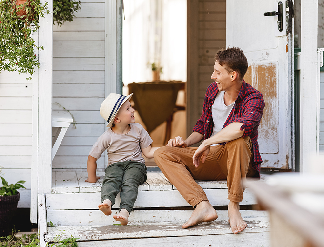 A barefoot man and a young boy wearing a hat sit on white wooden steps, smiling and holding hands while looking at each other.