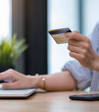 Person holding a credit card in one hand while using a laptop with the other hand at a wooden desk.