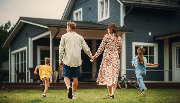 Parents holding hands walking toward a modern house while their two children run ahead on green grass.