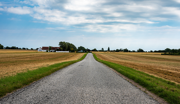 Straight rural road through dry harvested fields leading to a farmhouse under a partly cloudy blue sky.