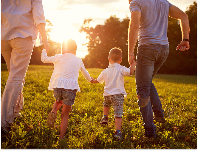 Two children holding hands with two adults walking through a sunlit grassy field at sunset.