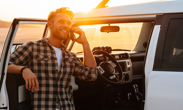 Smiling man in checkered shirt leaning on open door of a white car while talking on a phone during sunset.