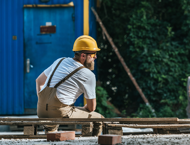 Construction worker wearing a yellow hard hat and beige overalls sitting on wooden pallets at a construction site.