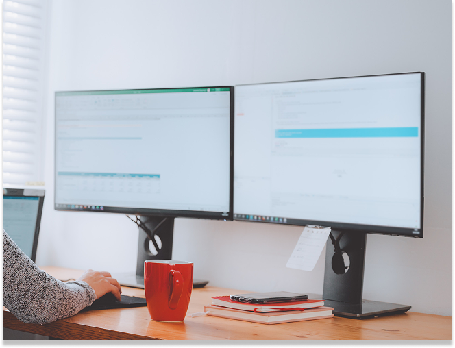 Person using a computer with dual monitors on a wooden desk with a red mug and stacked notebooks.