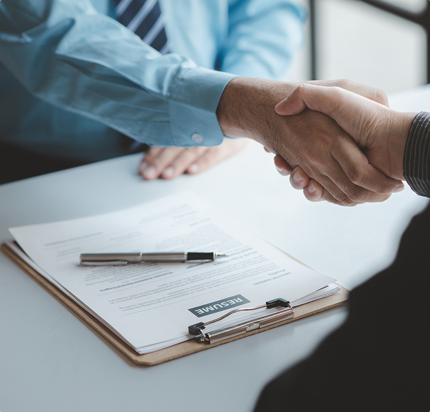 Two people shaking hands over a desk with a resume and pen on it.