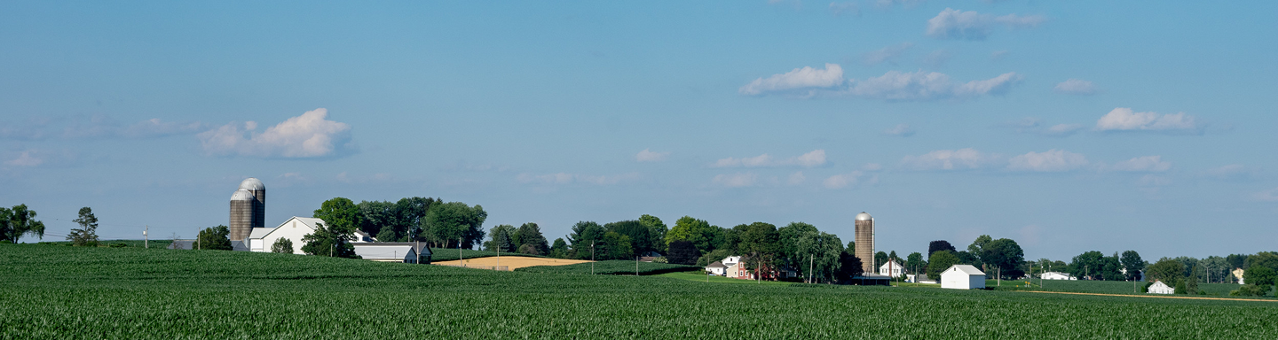 Wide farmland with green crops, white farm buildings, silos, and trees under a blue sky with scattered clouds.