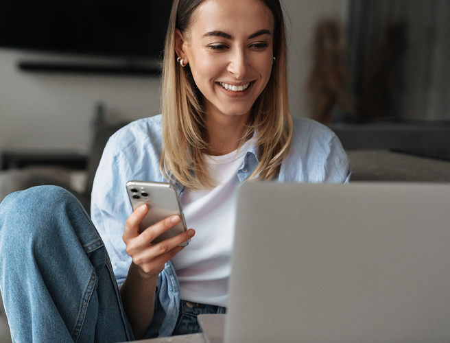 Smiling woman holding a smartphone and looking at a laptop screen indoors.