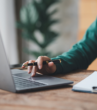 Person's hand holding a pen and using a laptop trackpad on a wooden desk.