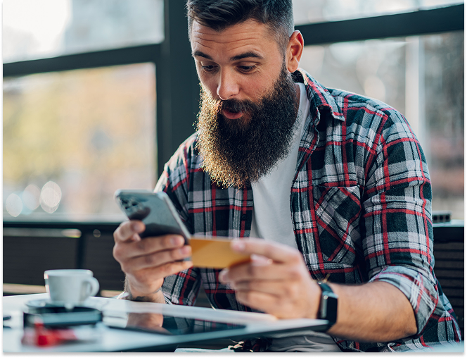 Bearded man in a plaid shirt looking surprised while holding a credit card and smartphone indoors.