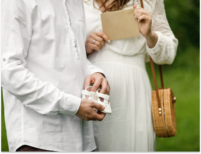 Man and woman in white clothing holding a baby shoe and a brown envelope outdoors.