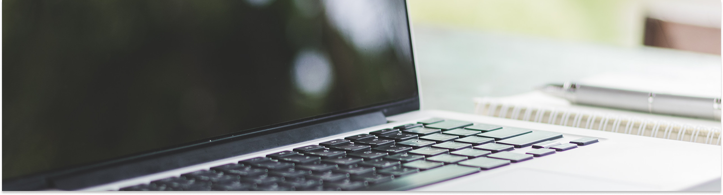 Close-up of a laptop keyboard and partially visible screen with a blurred notebook and pen in the background.