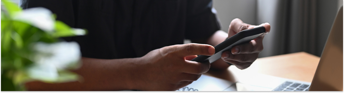 Person holding and using a smartphone at a desk with a laptop and a plant nearby.