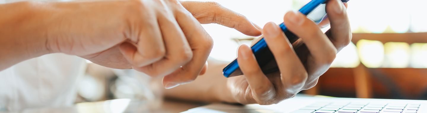 Close-up of hands holding and tapping a blue smartphone over a laptop keyboard.
