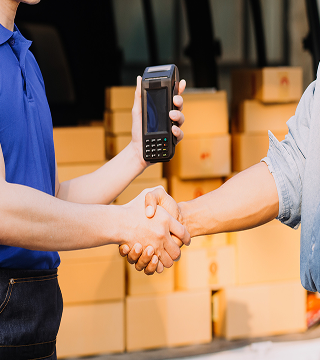 Two people shaking hands, one holding a payment terminal, with stacked cardboard boxes in the background.