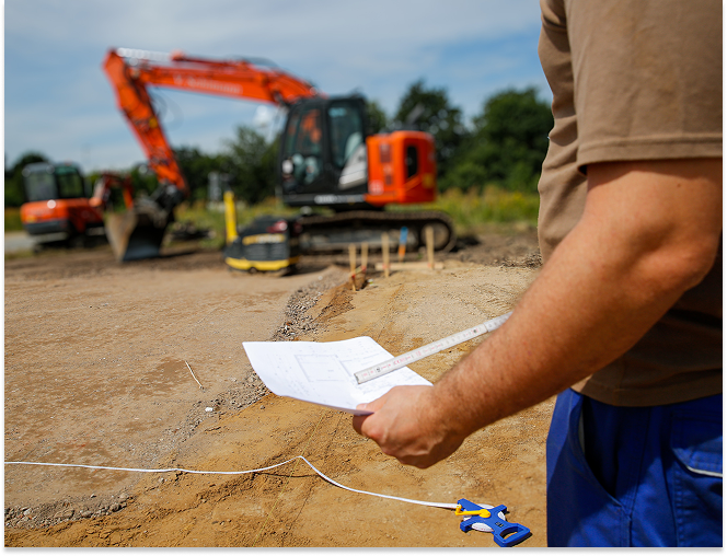 Construction worker holding blueprints and measuring tape at a dirt site with an orange excavator in the background.