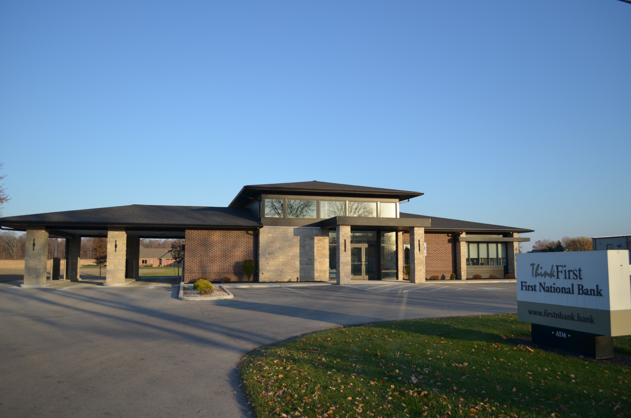 Building entrance with a large pine tree to the left and concrete walkway leading to glass doors under a dark sloped roof.