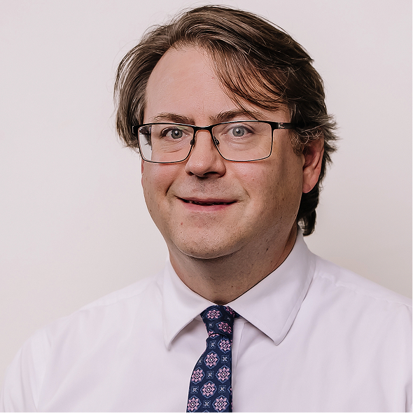 Man with brown hair and glasses wearing a dark suit, blue shirt, and striped tie, smiling against a blurred brown background.