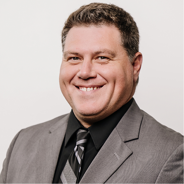 Professional headshot of a man with short dark hair wearing a black suit, white shirt, and patterned tie against a neutral background.