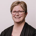 Smiling woman with short brown hair, black glasses, and a blue top against a gray background.
