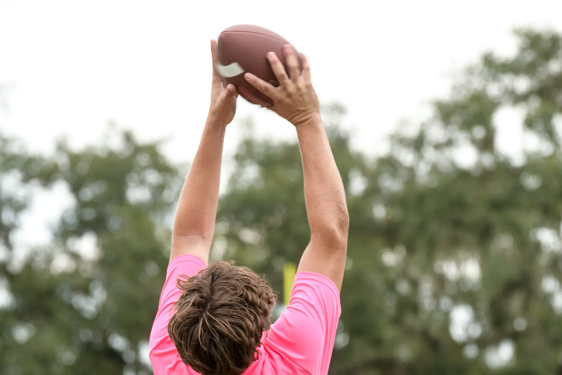 A young athlete in a pink shirt reaches up to catch a football against a backdrop of trees and cloudy skies.