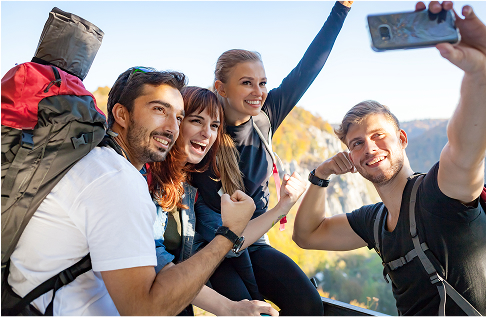 A group of hikers with backpacks posing for a selfie on a mountain trail, celebrating and flexing their arms with scenic views in the background.