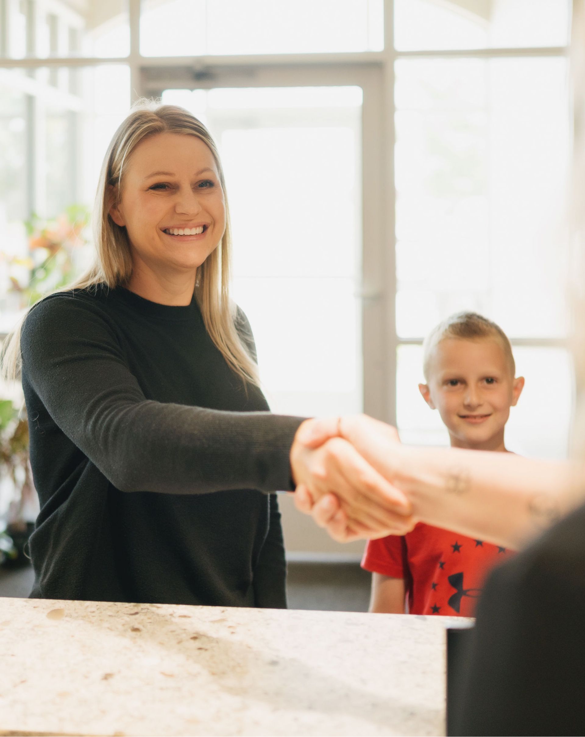 Smiling woman shaking hands with another person, with a young boy smiling in the background.