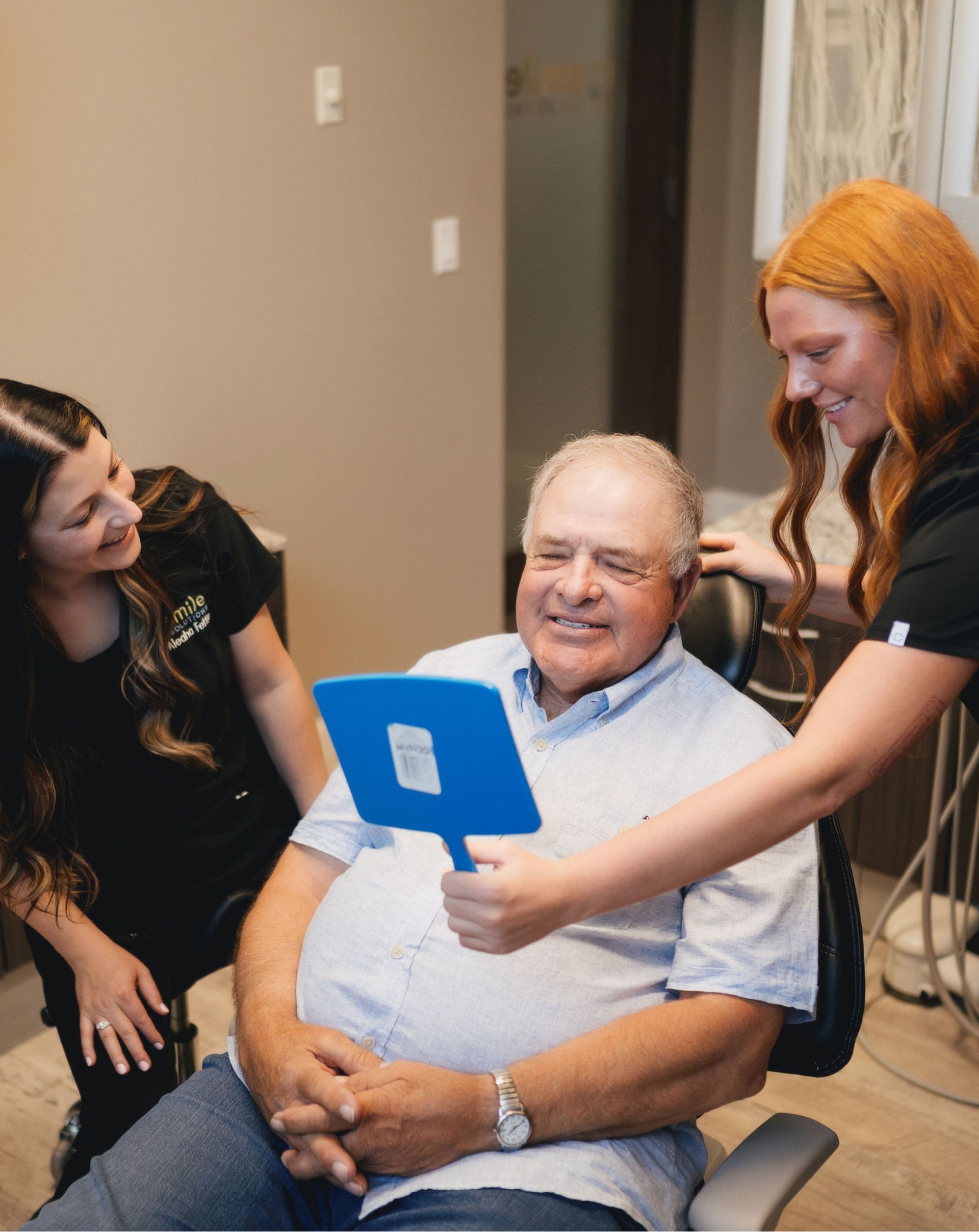 Elderly man sitting in a dental chair smiling while two female dental professionals show him his reflection in a blue hand mirror.