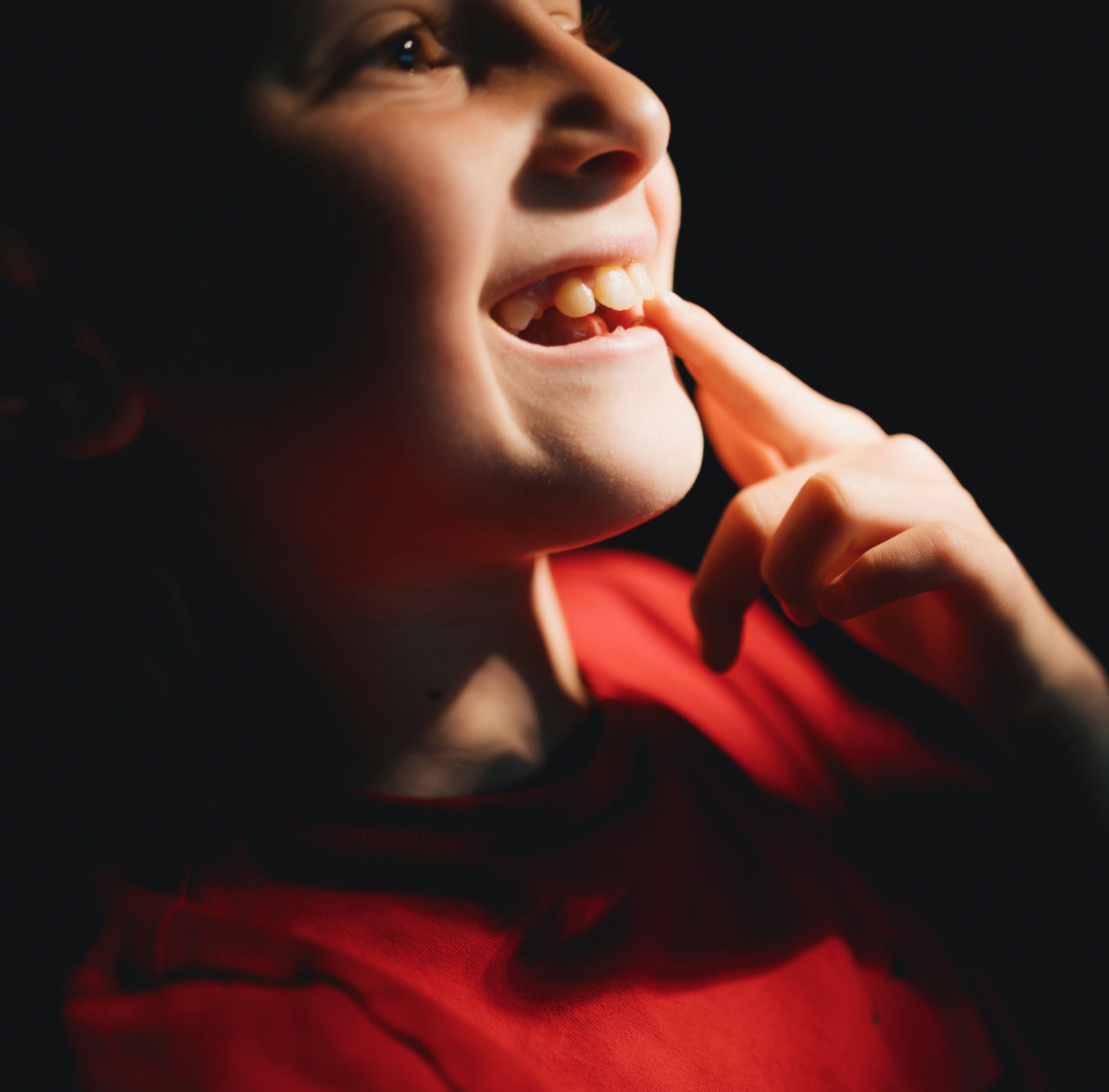 Close-up of a child smiling and pointing at a missing tooth while wearing a red shirt.