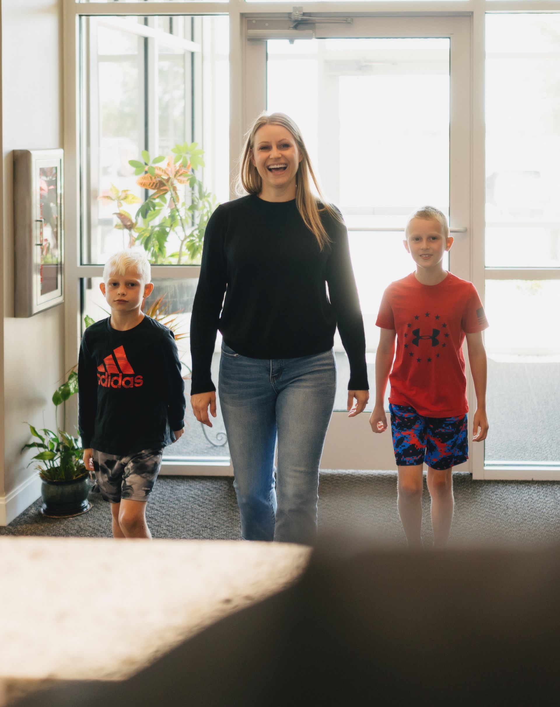 Smiling woman walking indoors with two young boys, one wearing a black Adidas shirt and the other a red Under Armour shirt.