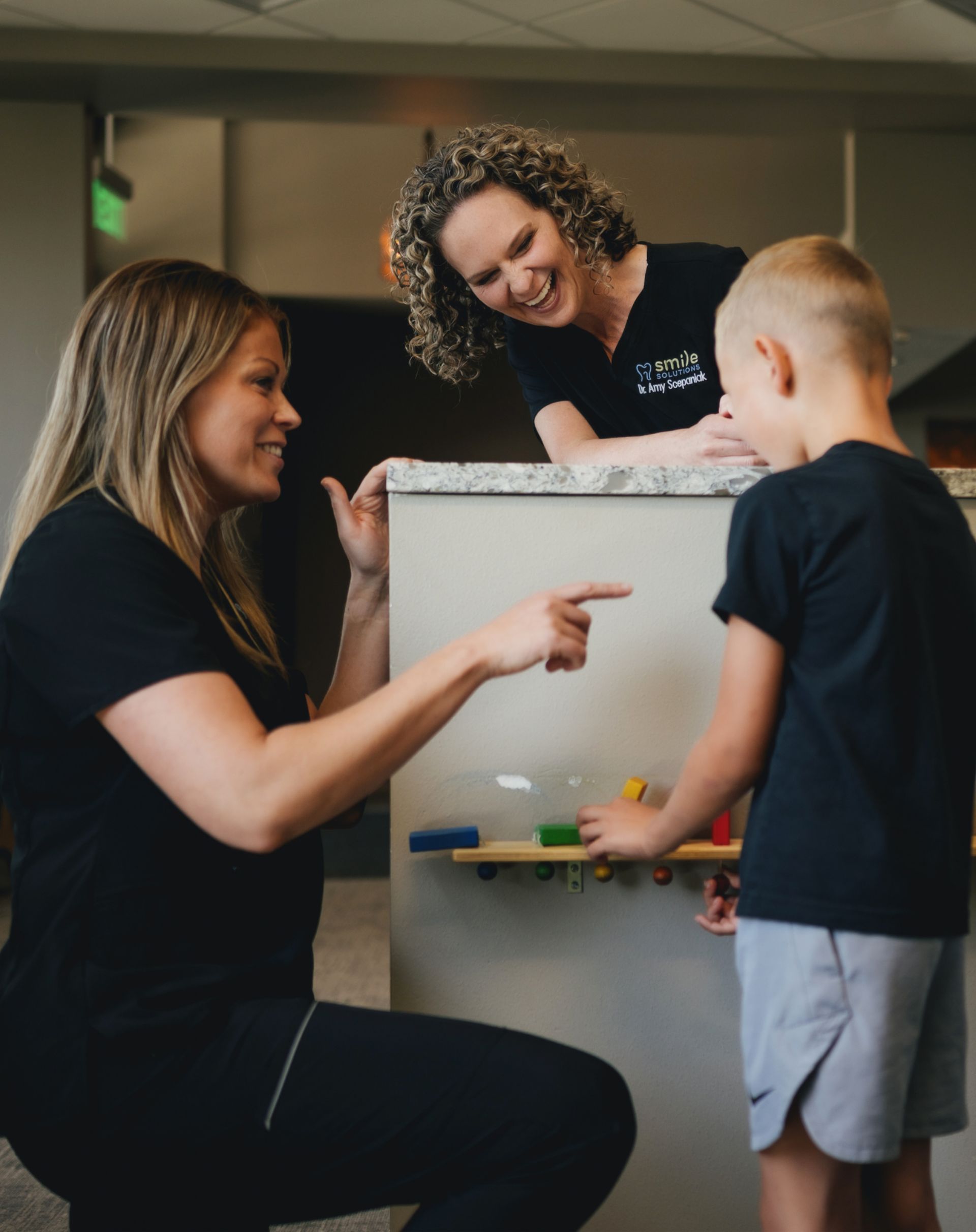 Two women in black scrubs interacting with a young boy playing with colorful wooden blocks on a wall-mounted shelf.