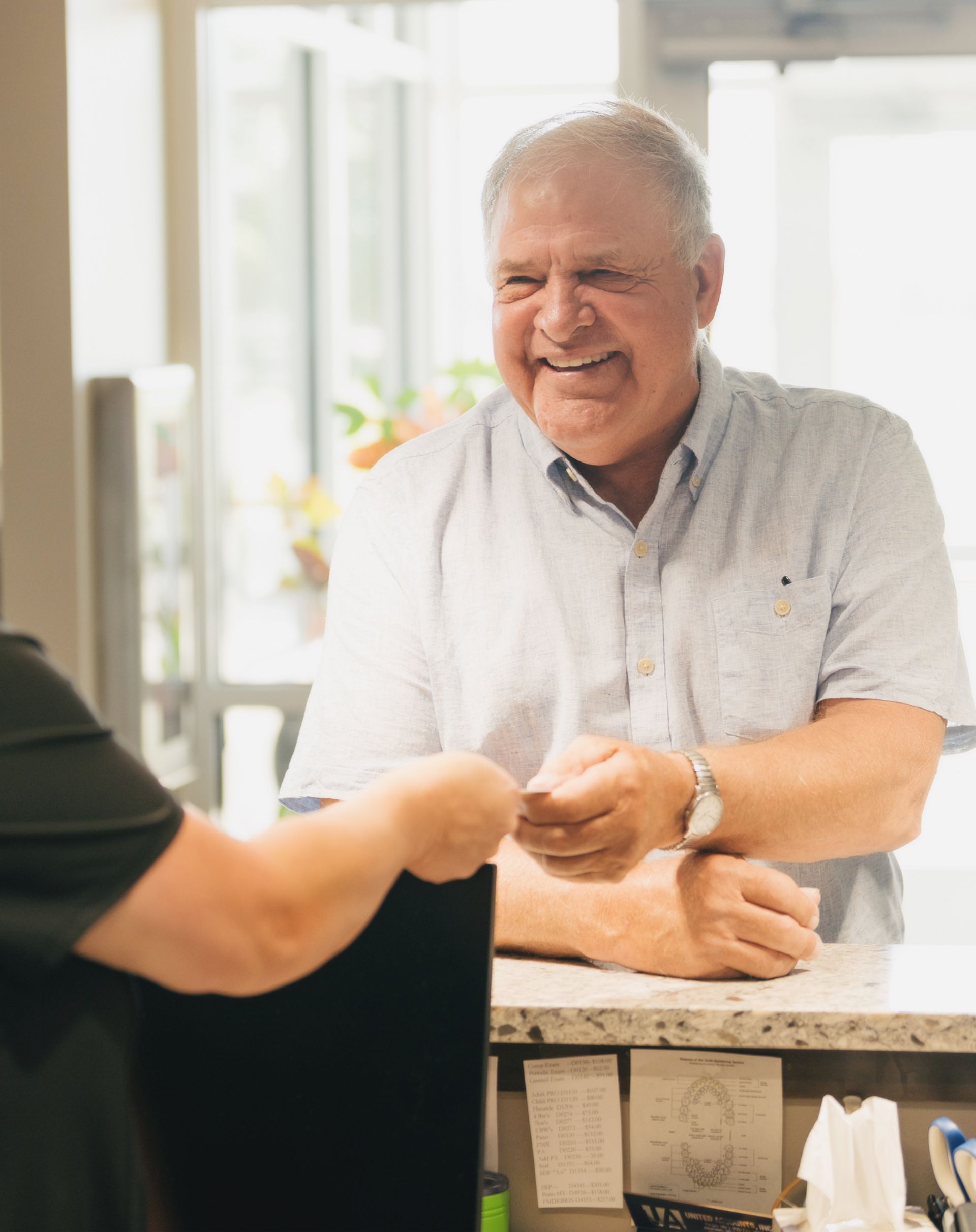 Smiling elderly man handing a card to another person across a reception desk in a bright room.
