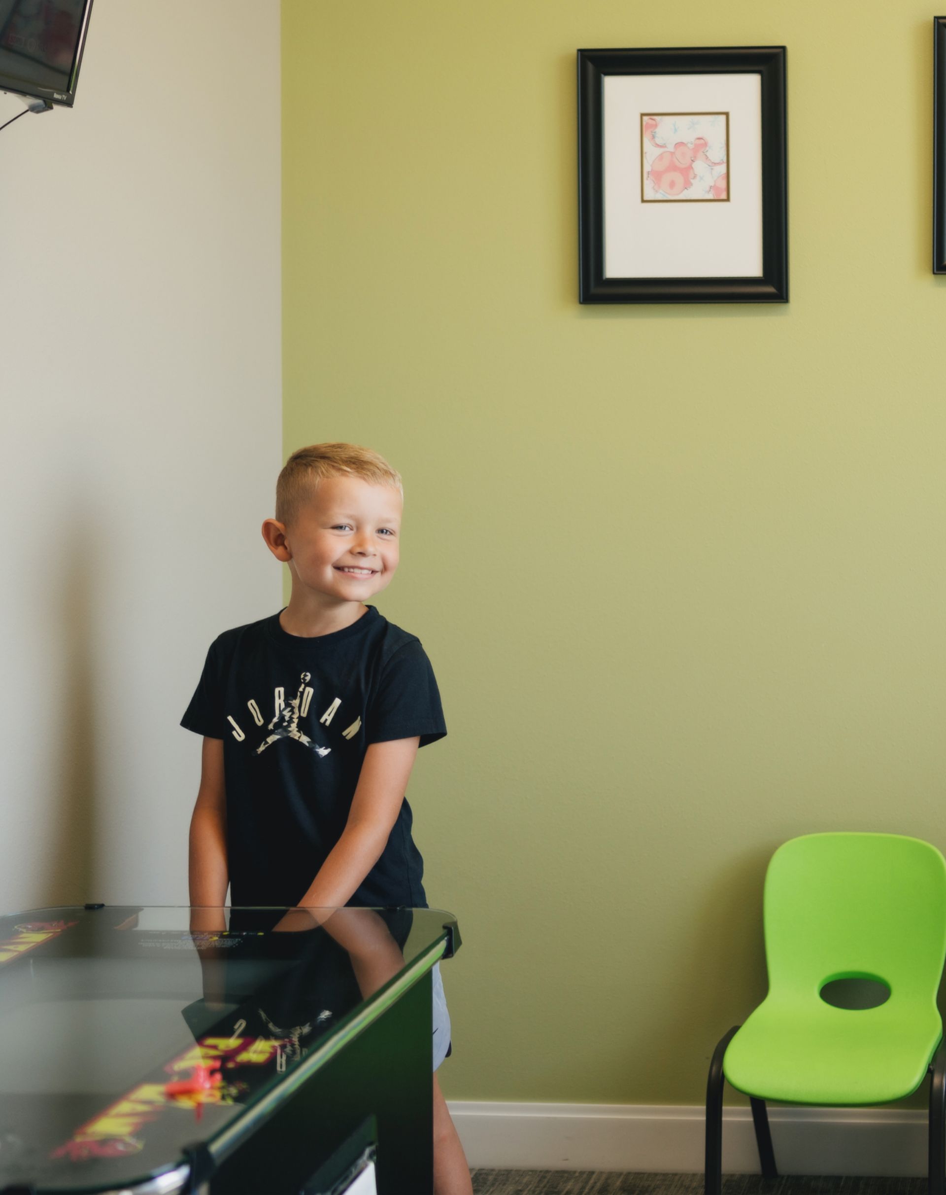 Smiling young boy in a black Jordan t-shirt standing next to a game machine in a room with light green walls and a green chair.