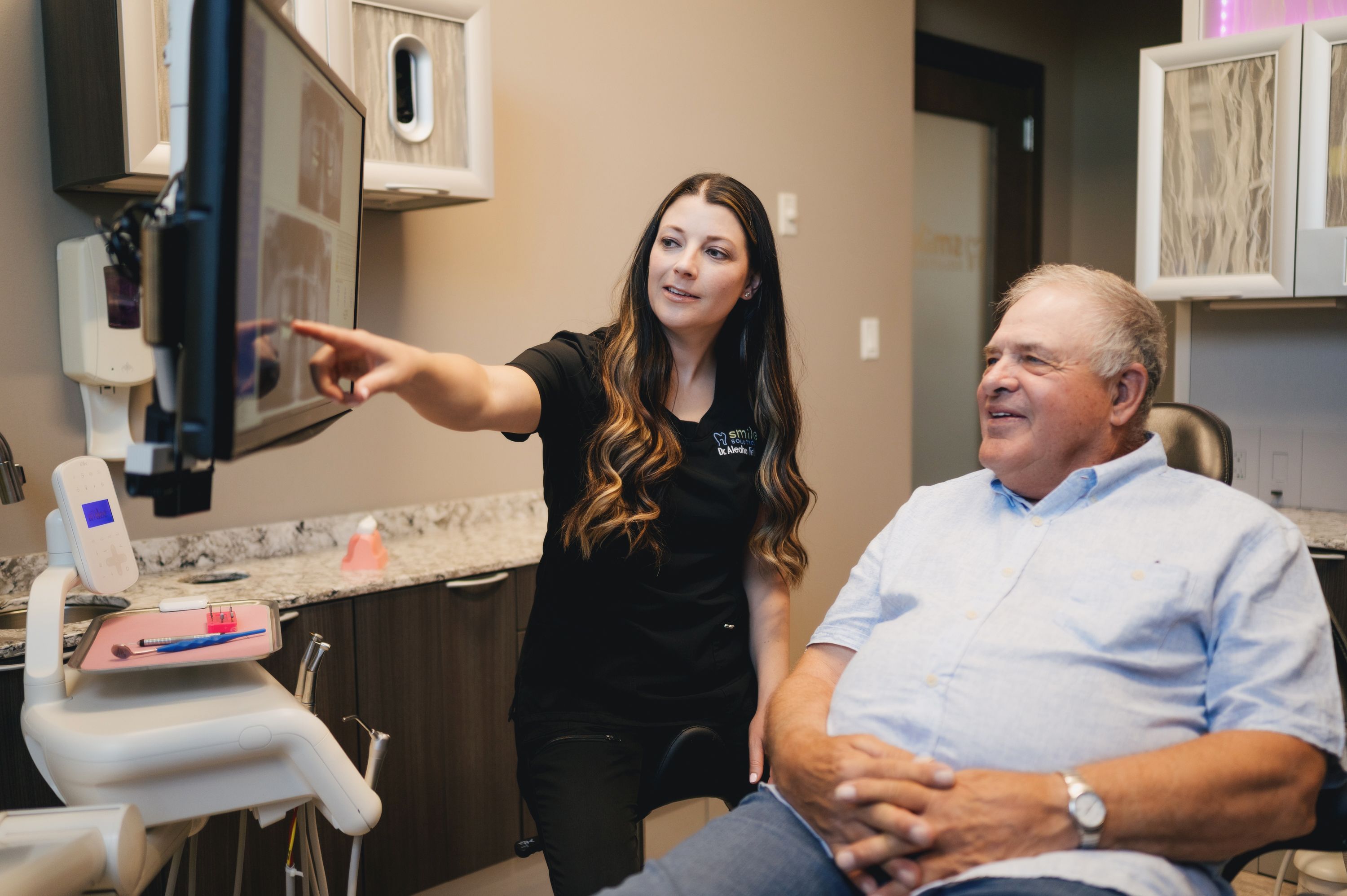 Dentist explaining dental X-rays on a screen to an elderly male patient sitting in a dental chair.