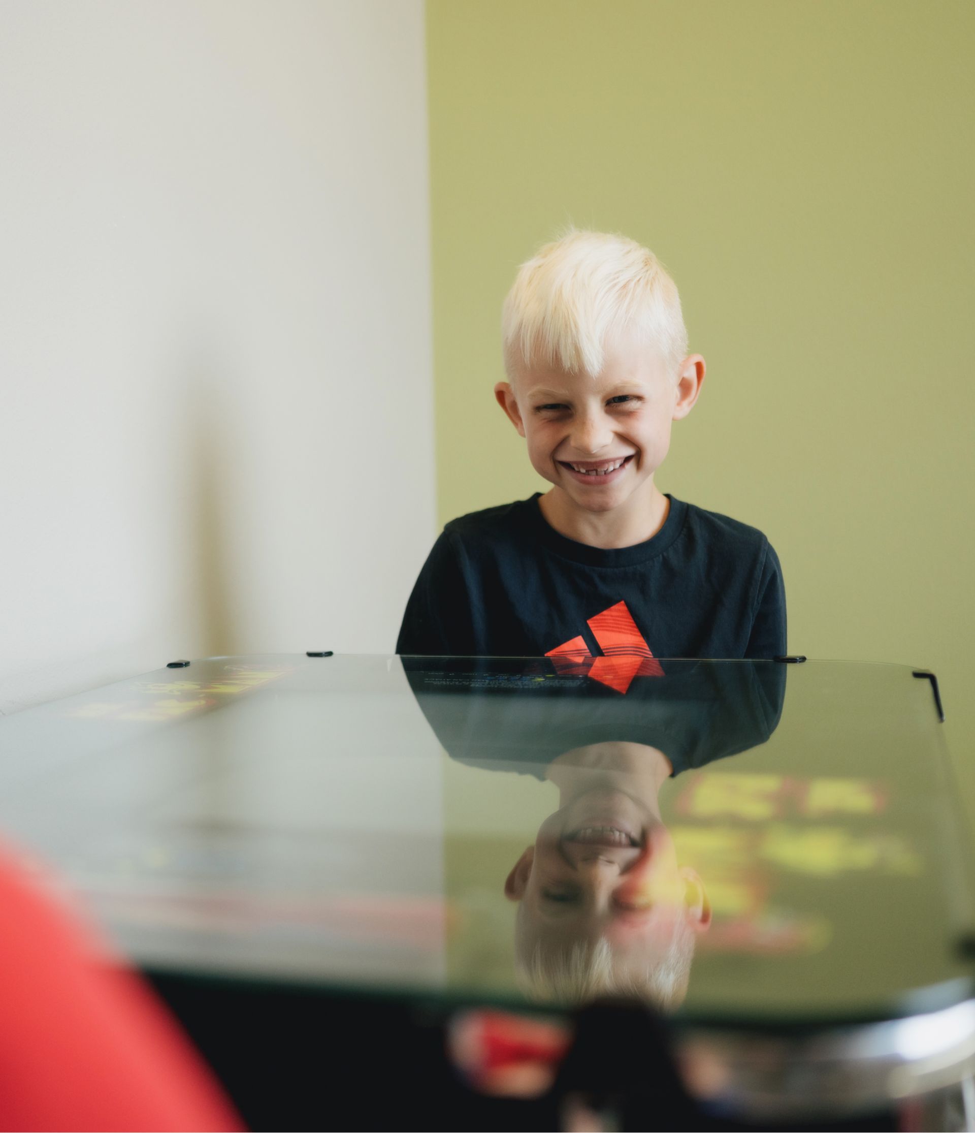 Smiling blonde boy wearing a black shirt with a red logo, reflected on a glass surface in front of him.
