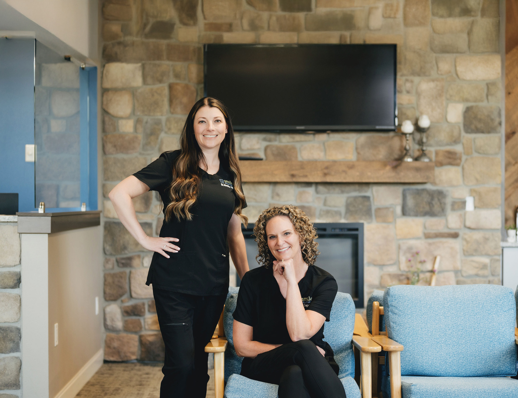 Two smiling women in black scrubs pose in a cozy waiting area with stone walls and a TV mounted above a fireplace.