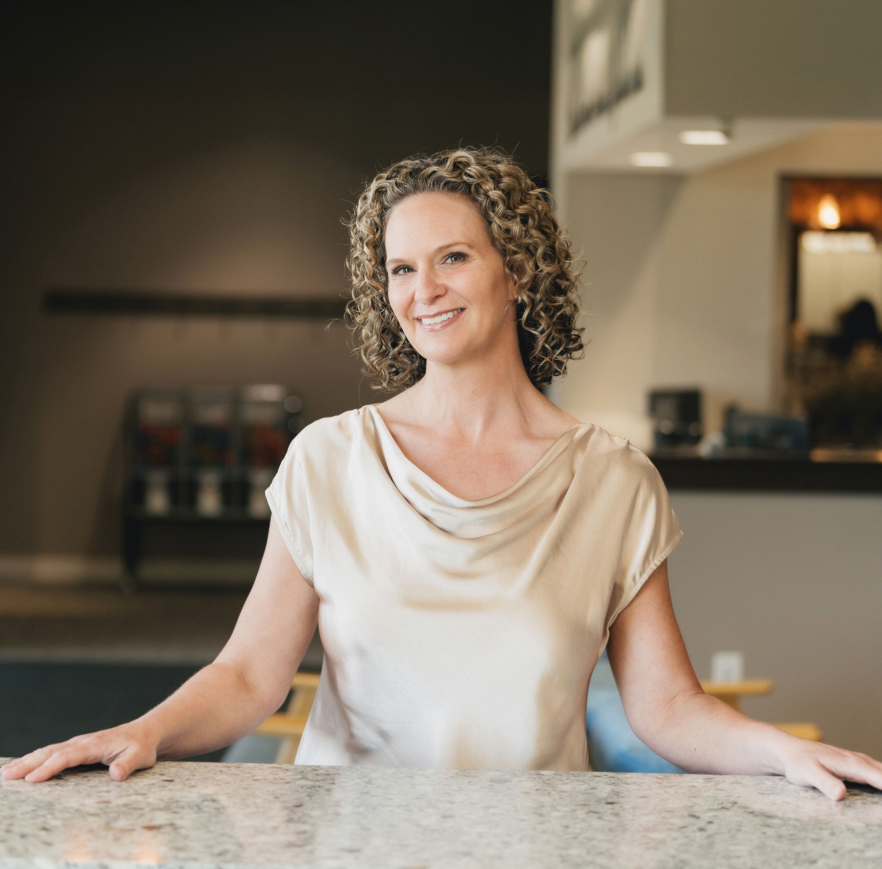 Smiling woman with curly hair in a cream blouse standing behind a marble countertop in a warmly lit room.