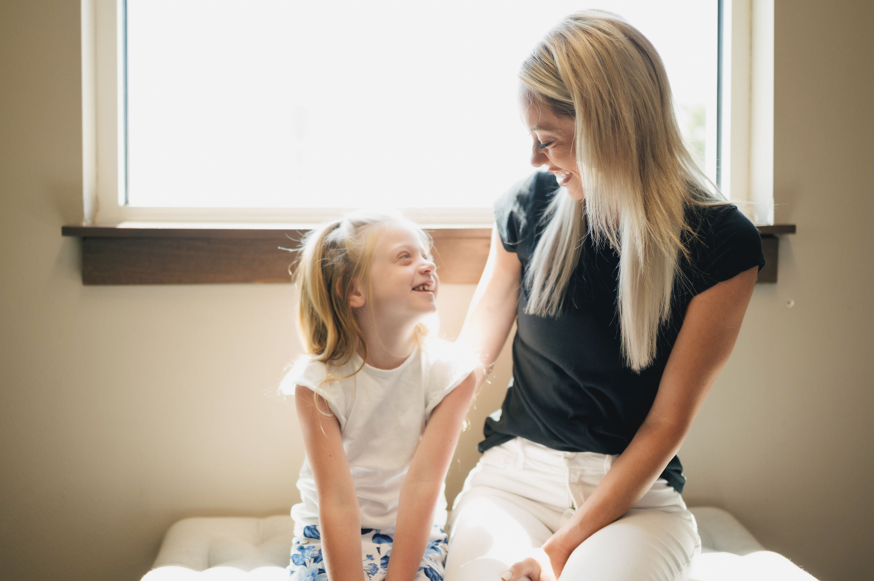 Smiling woman and young girl sitting together by a window, looking at each other warmly.