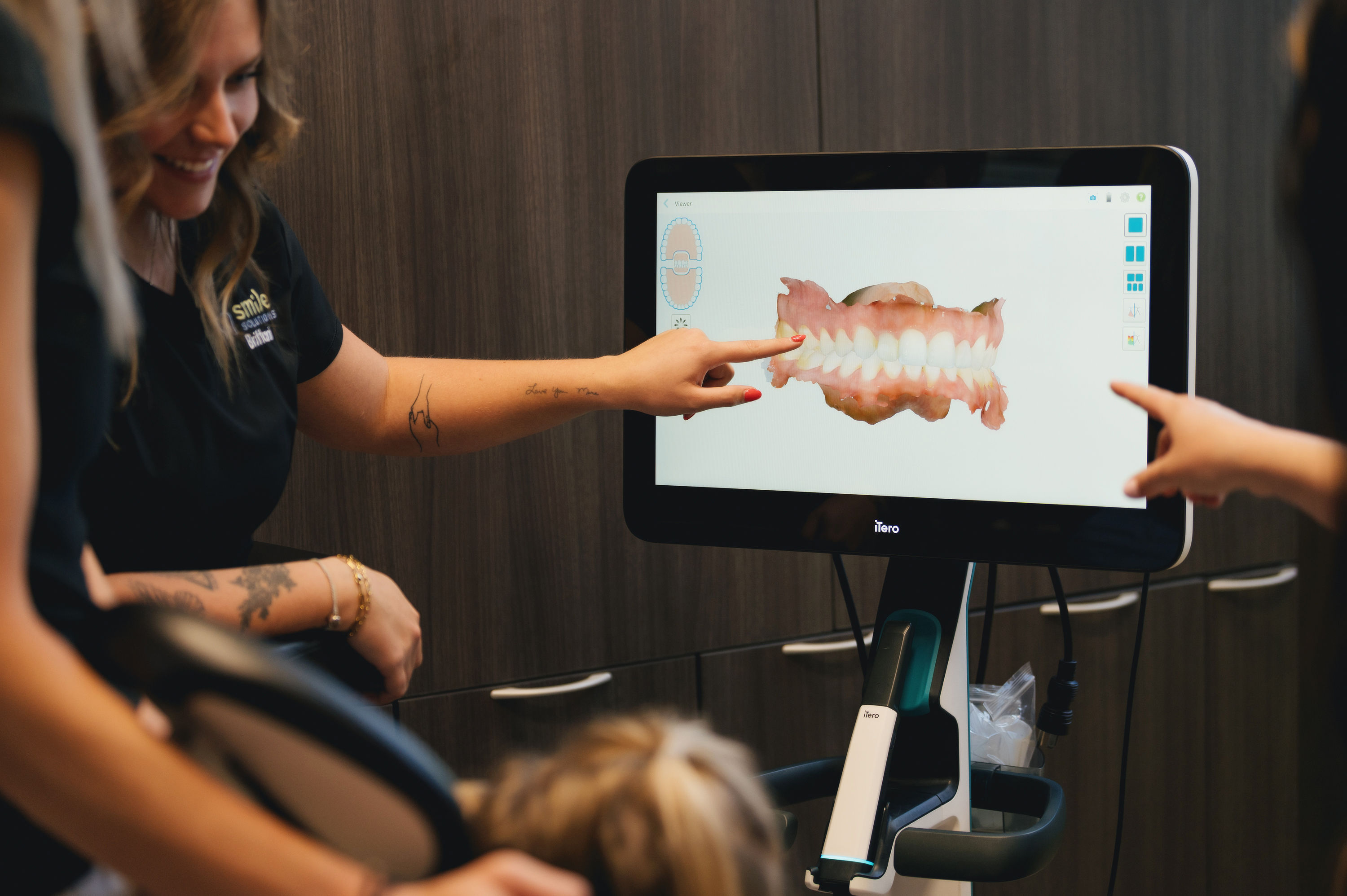 Dental professional pointing at a 3D digital scan of a patient's teeth on a screen in a dental office.