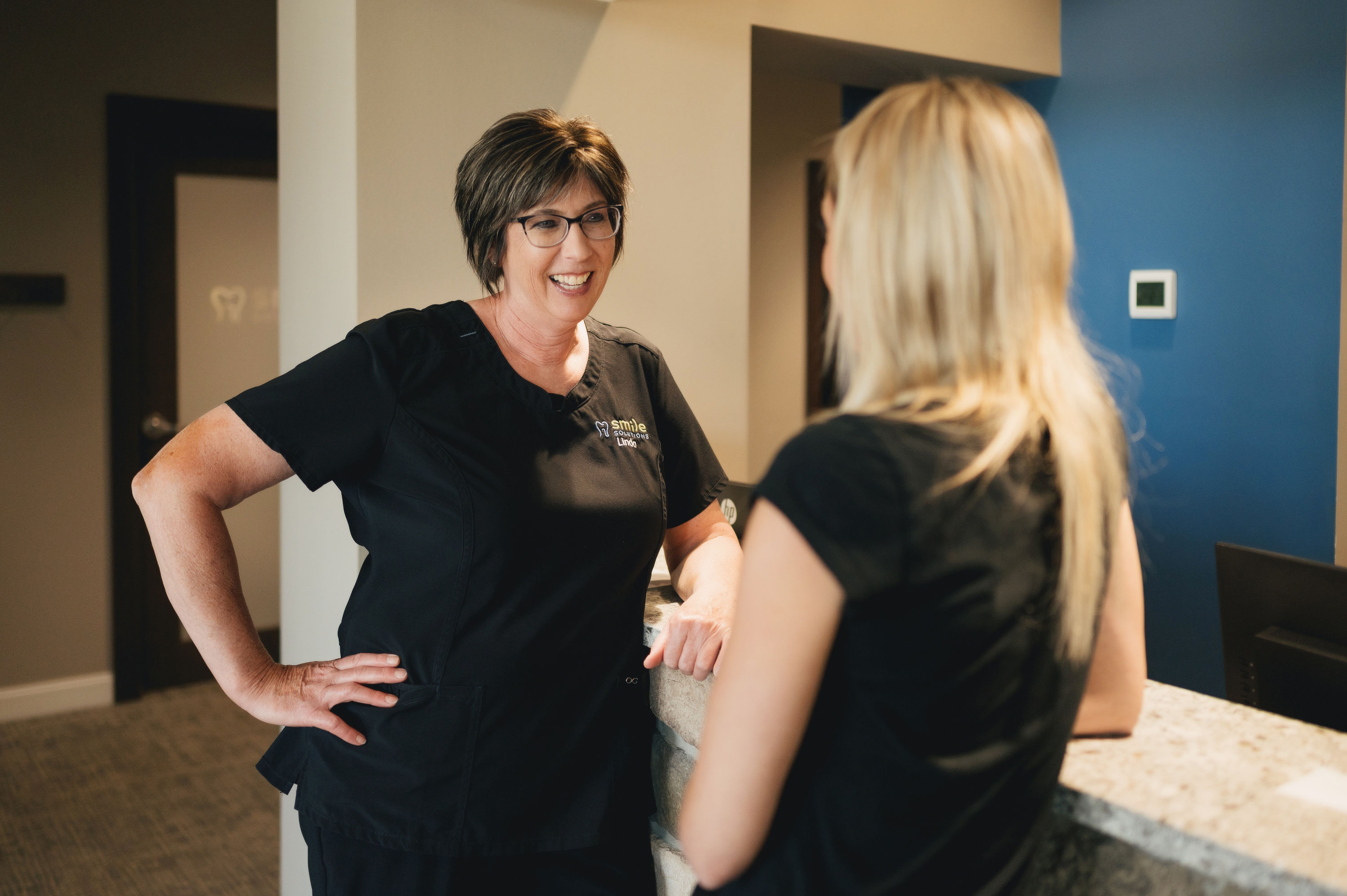 Smiling female dental professional in black scrubs talking to a blonde woman at the reception desk.