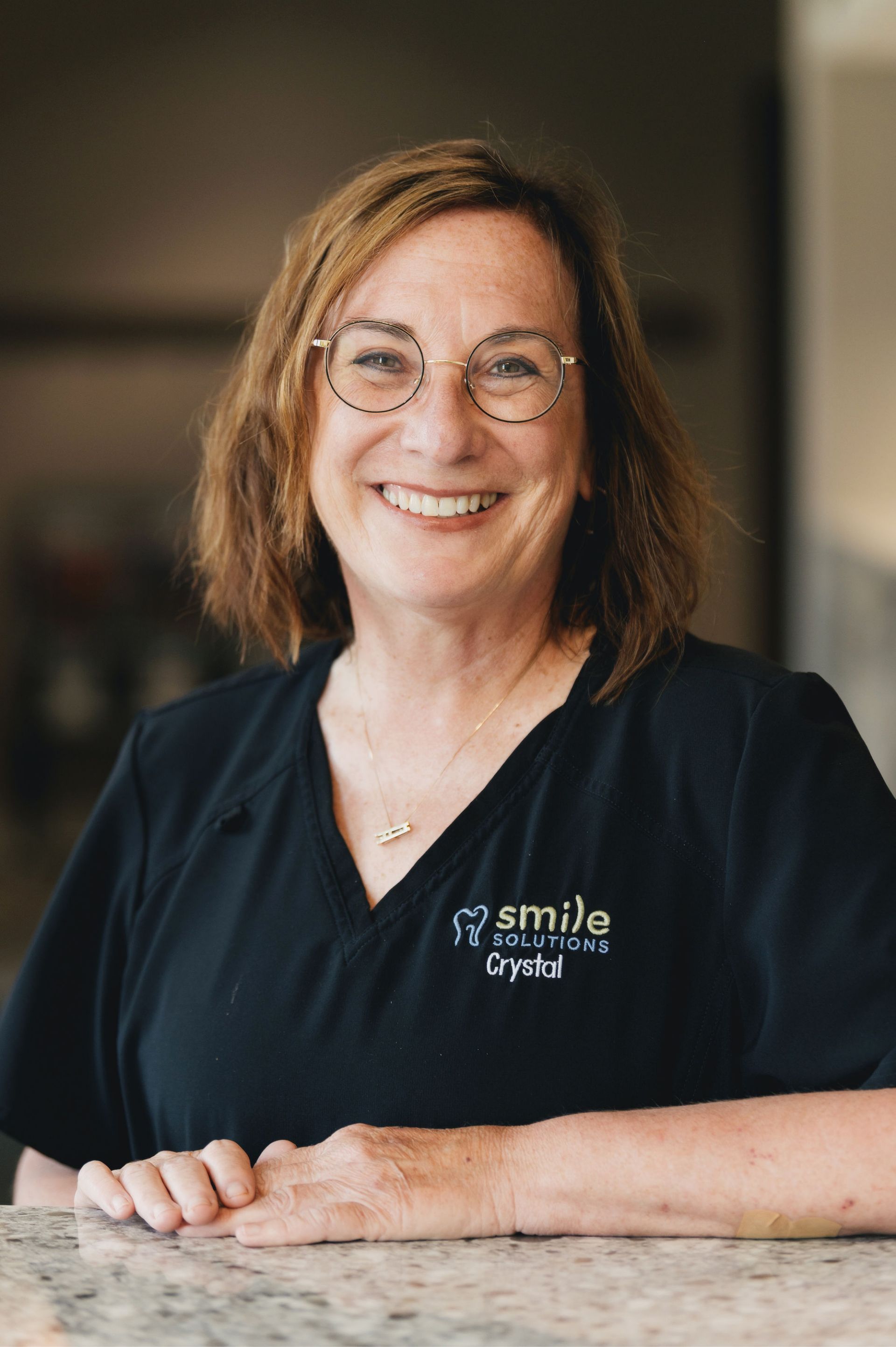 Smiling woman with glasses wearing a black shirt embroidered with 'smile solutions Crystal' resting her hands on a granite countertop.