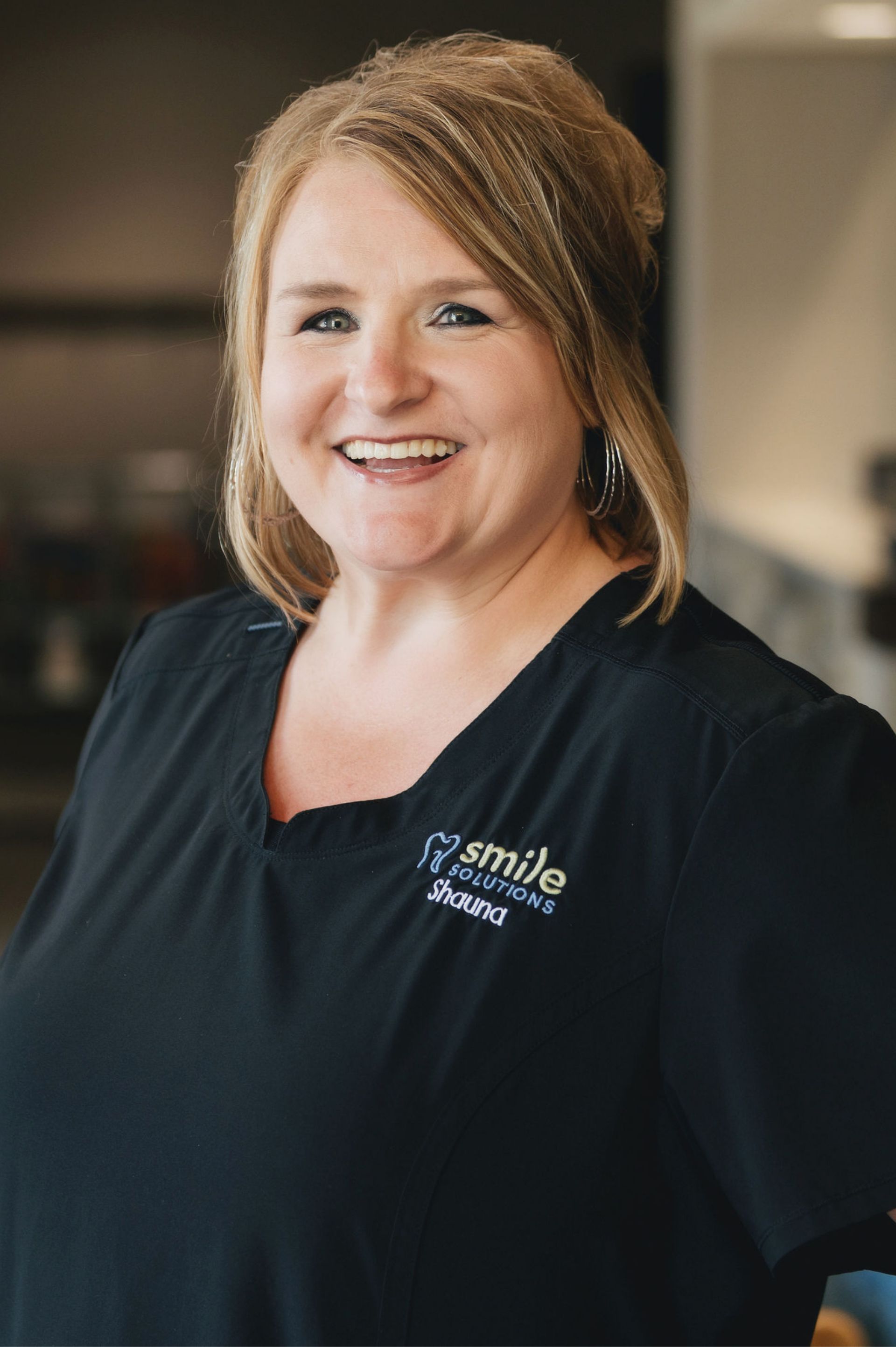 Smiling woman with blonde hair wearing a black shirt embroidered with 'Smile Solutions Shauna'.