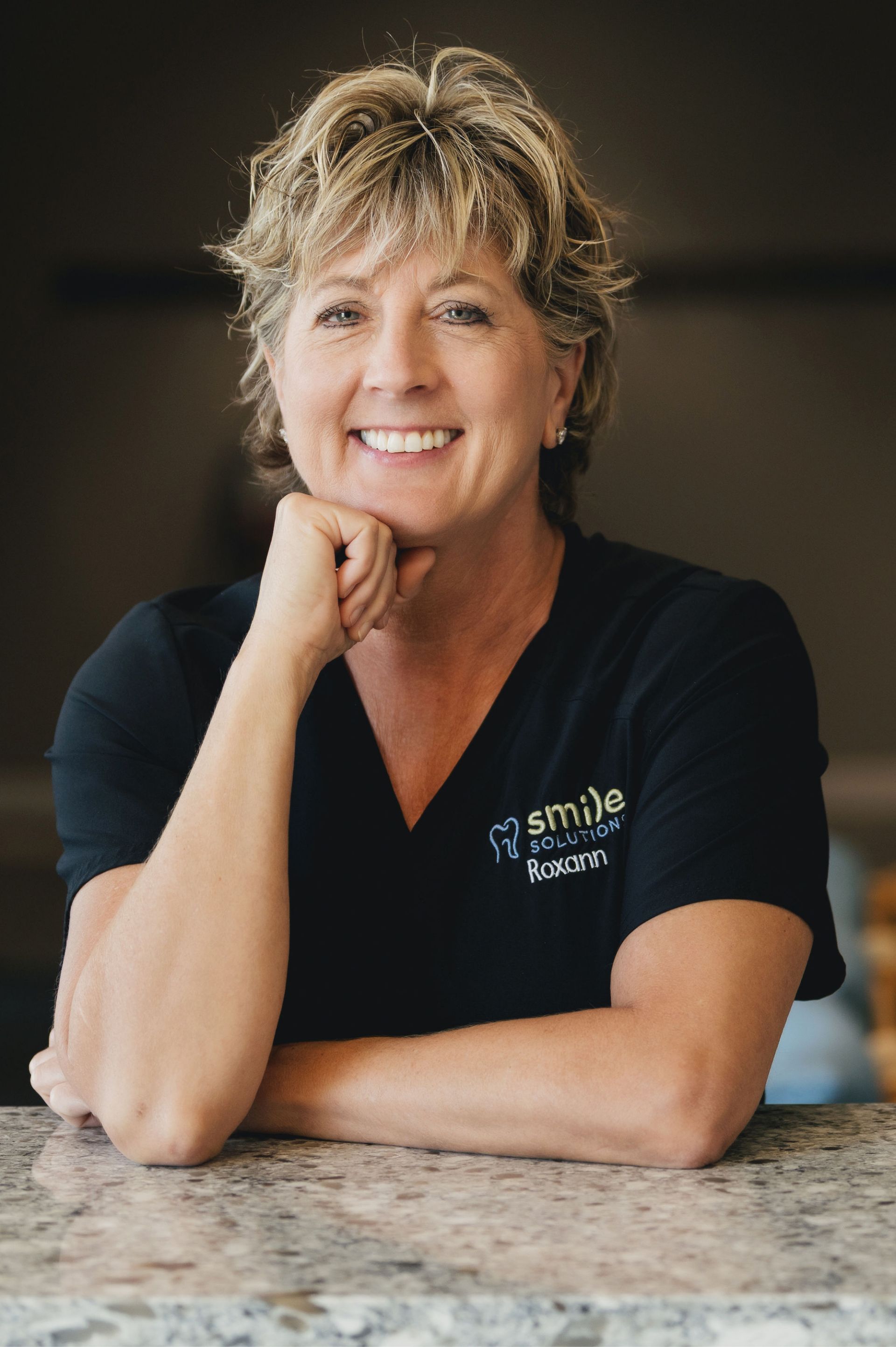 Smiling woman with short blonde hair wearing a black shirt with 'Smile Solutions Roxann' embroidered, resting her chin on her hand at a granite counter.
