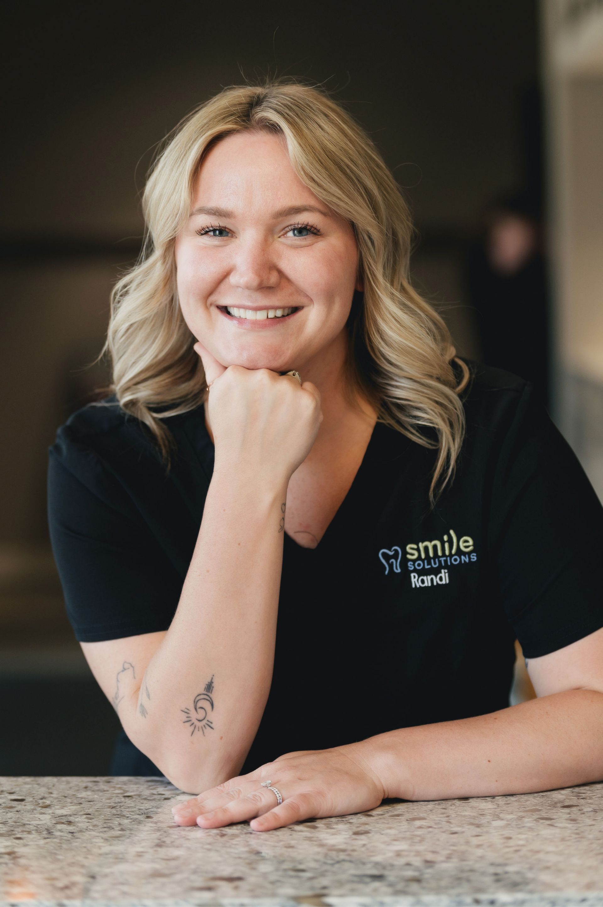 Smiling woman with blonde hair resting her chin on her hand wearing a black shirt with Smile Solutions logo and the name Randi.