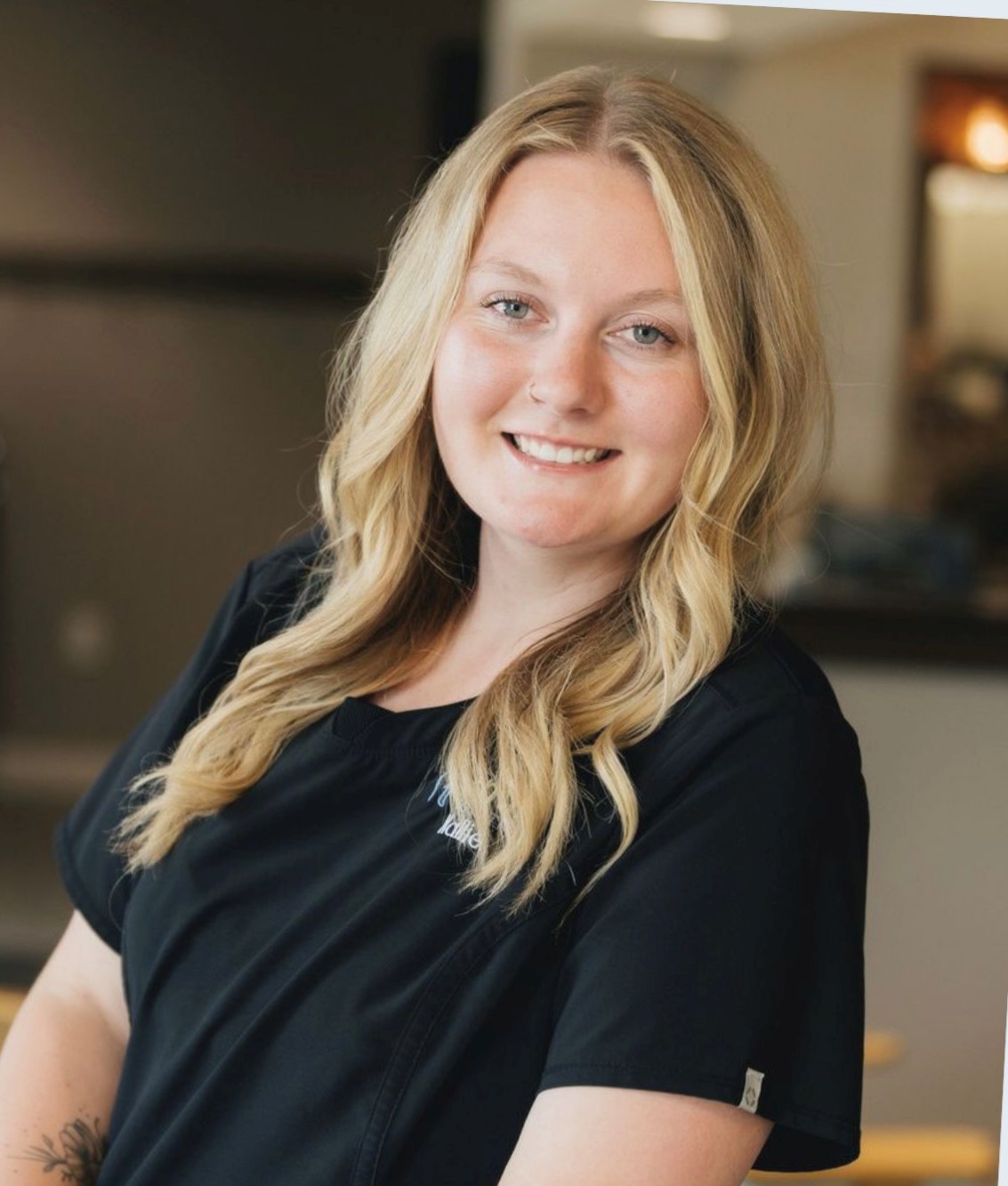 Smiling woman with long blonde hair wearing a black top in an indoor setting.