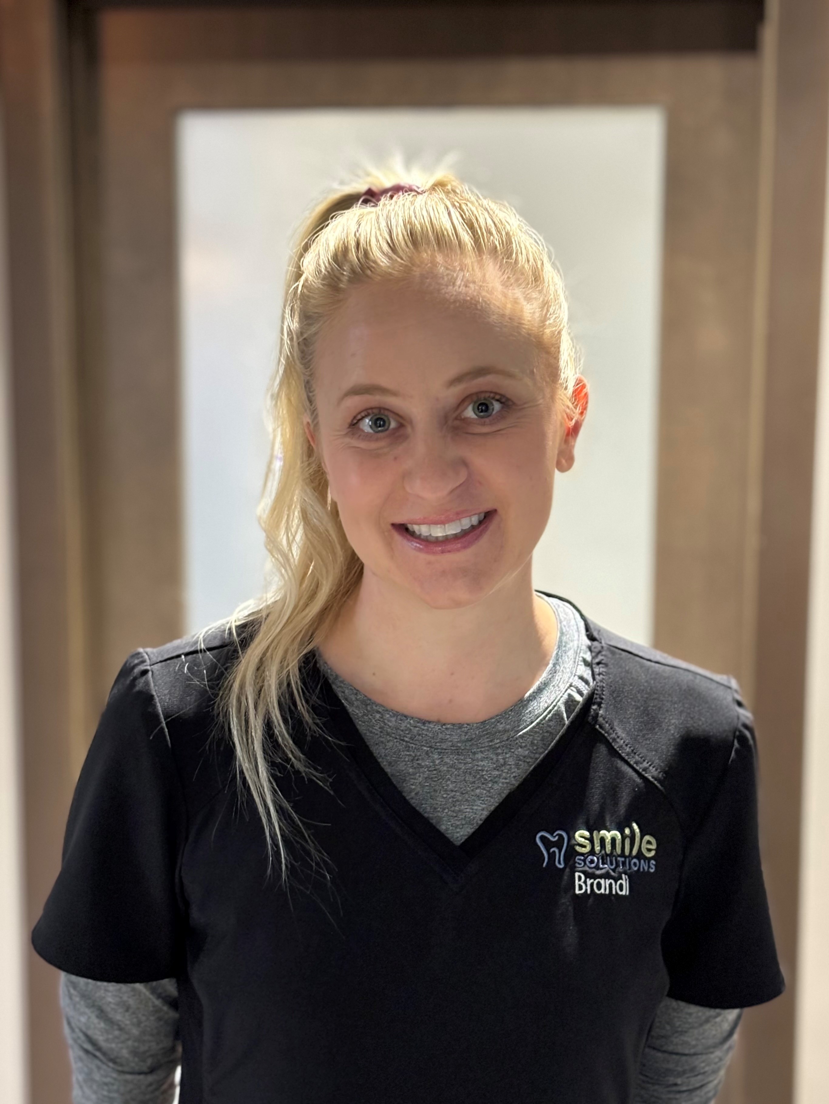 Smiling woman with blonde hair tied back, wearing black scrubs embroidered with 'smile SOLUTIONS Brandi'.