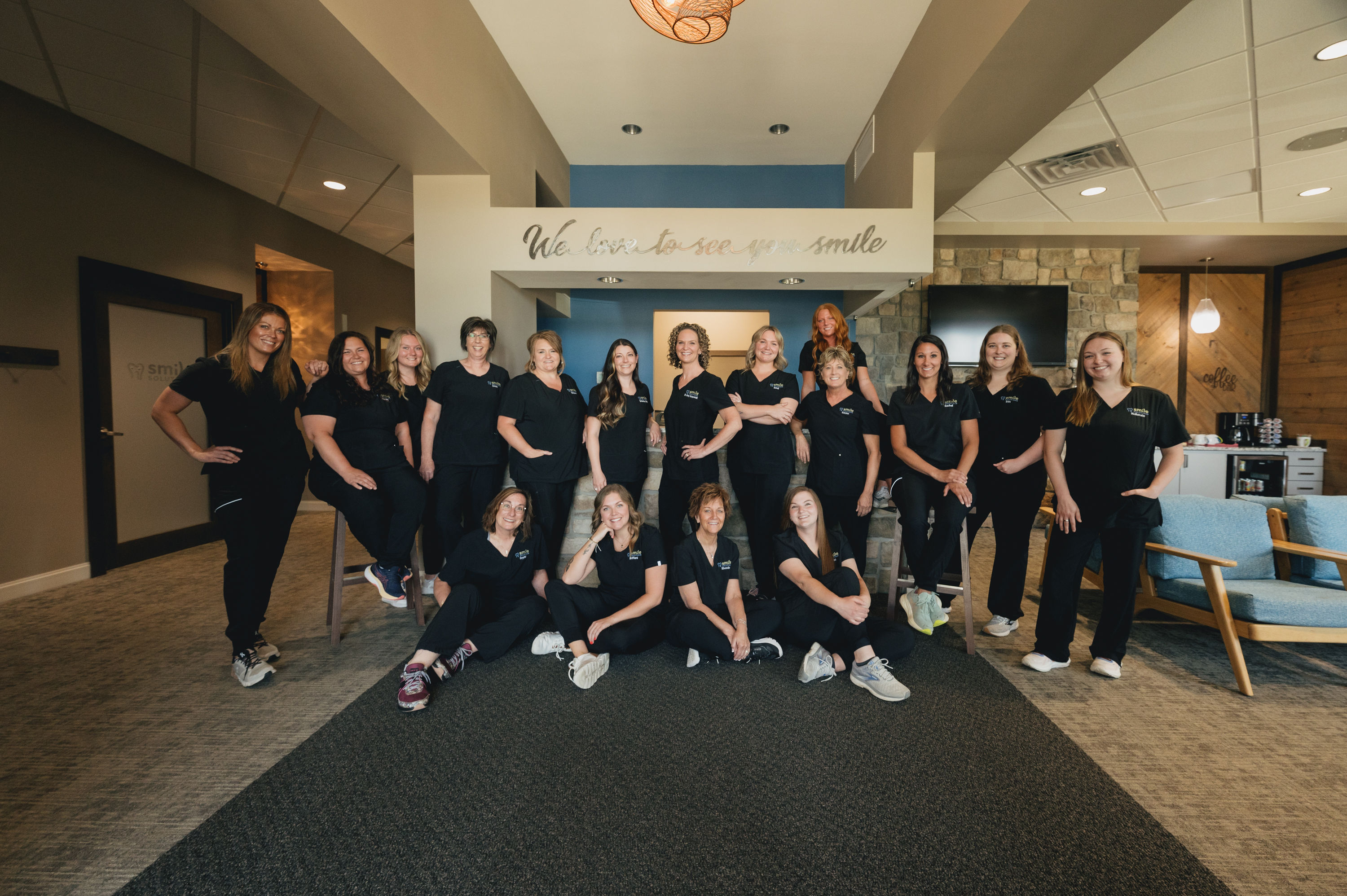 Group of 17 women in black uniforms posing together inside a modern office with a sign reading 'We love to see you smile'.