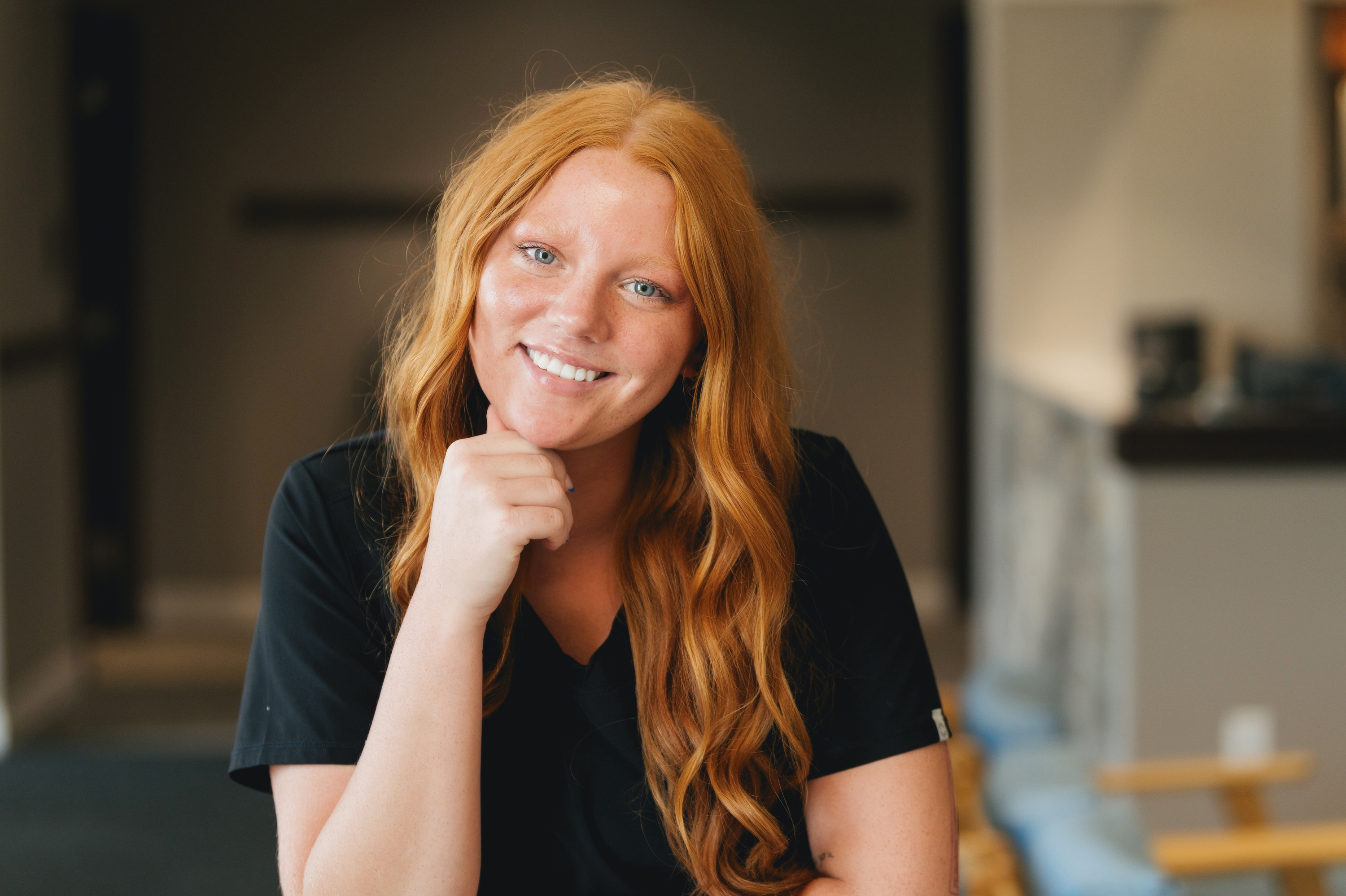 Smiling red-haired woman in black shirt resting her chin on her hand in a softly lit indoor setting.