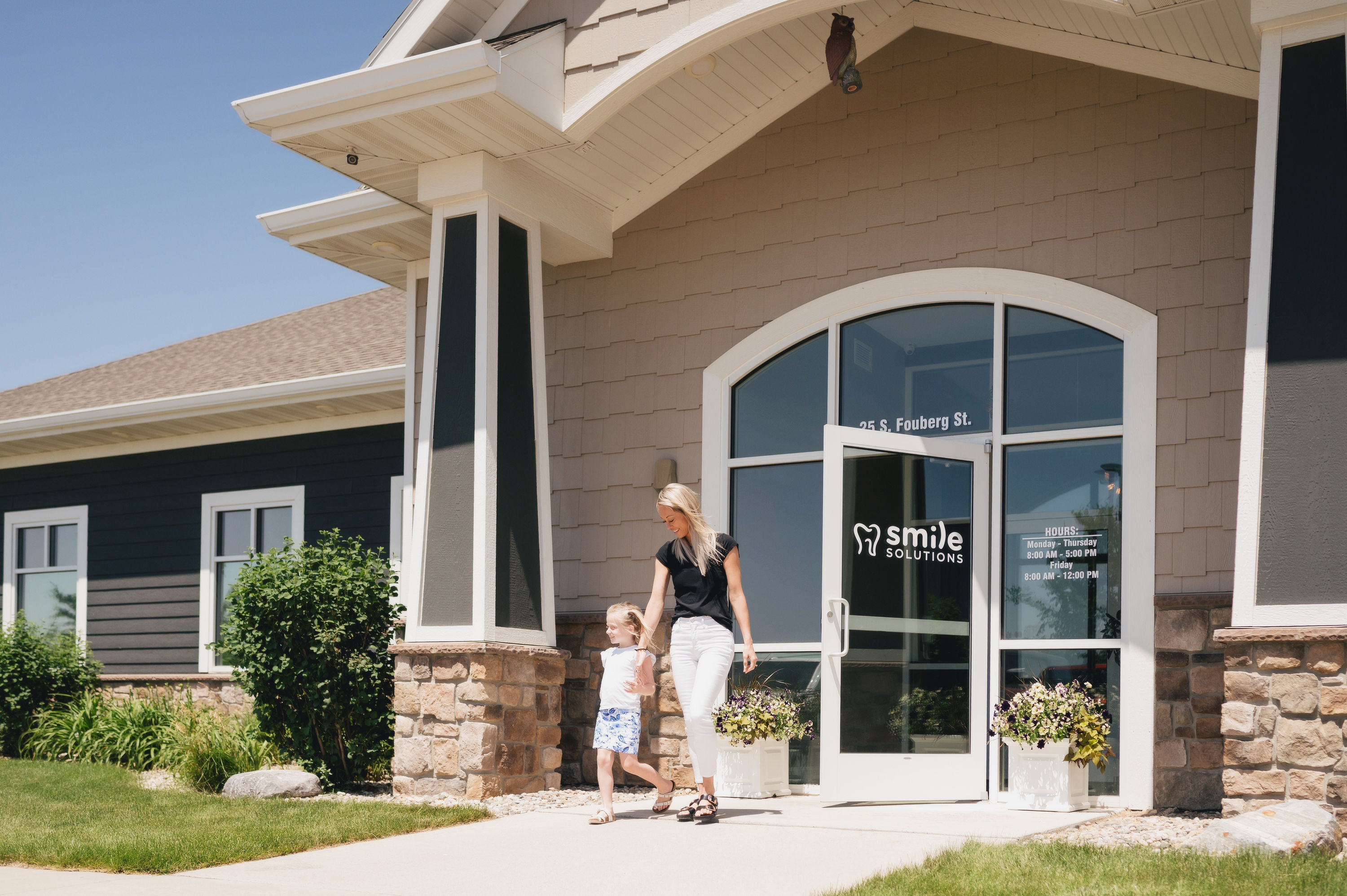A woman and a young girl walking hand in hand outside the entrance of Smile Solutions dental office on a sunny day.