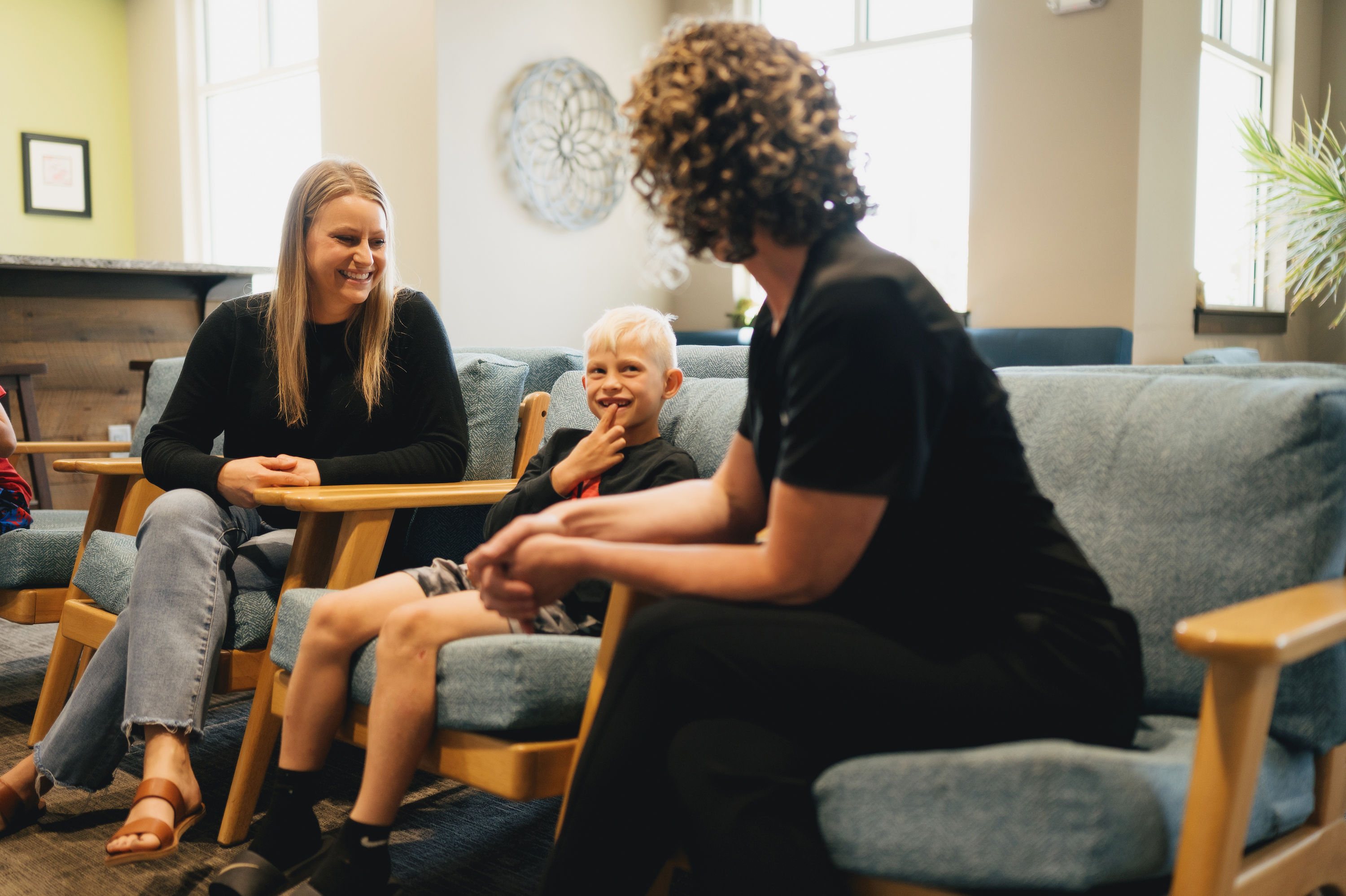 Young boy smiling and sitting between two women on blue cushioned wooden chairs in a well-lit room.