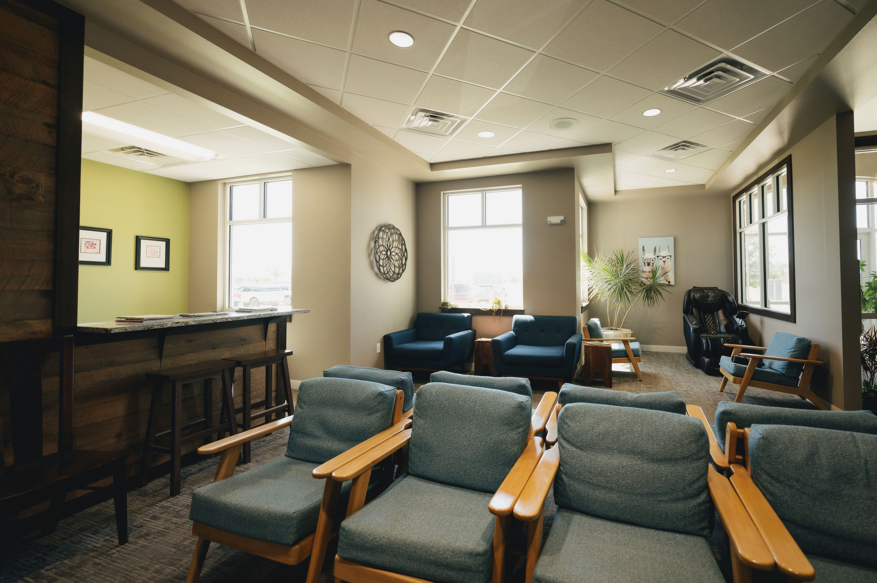 Well-lit waiting room with multiple blue cushioned wooden chairs, a reception desk, two large windows, and a massage chair.